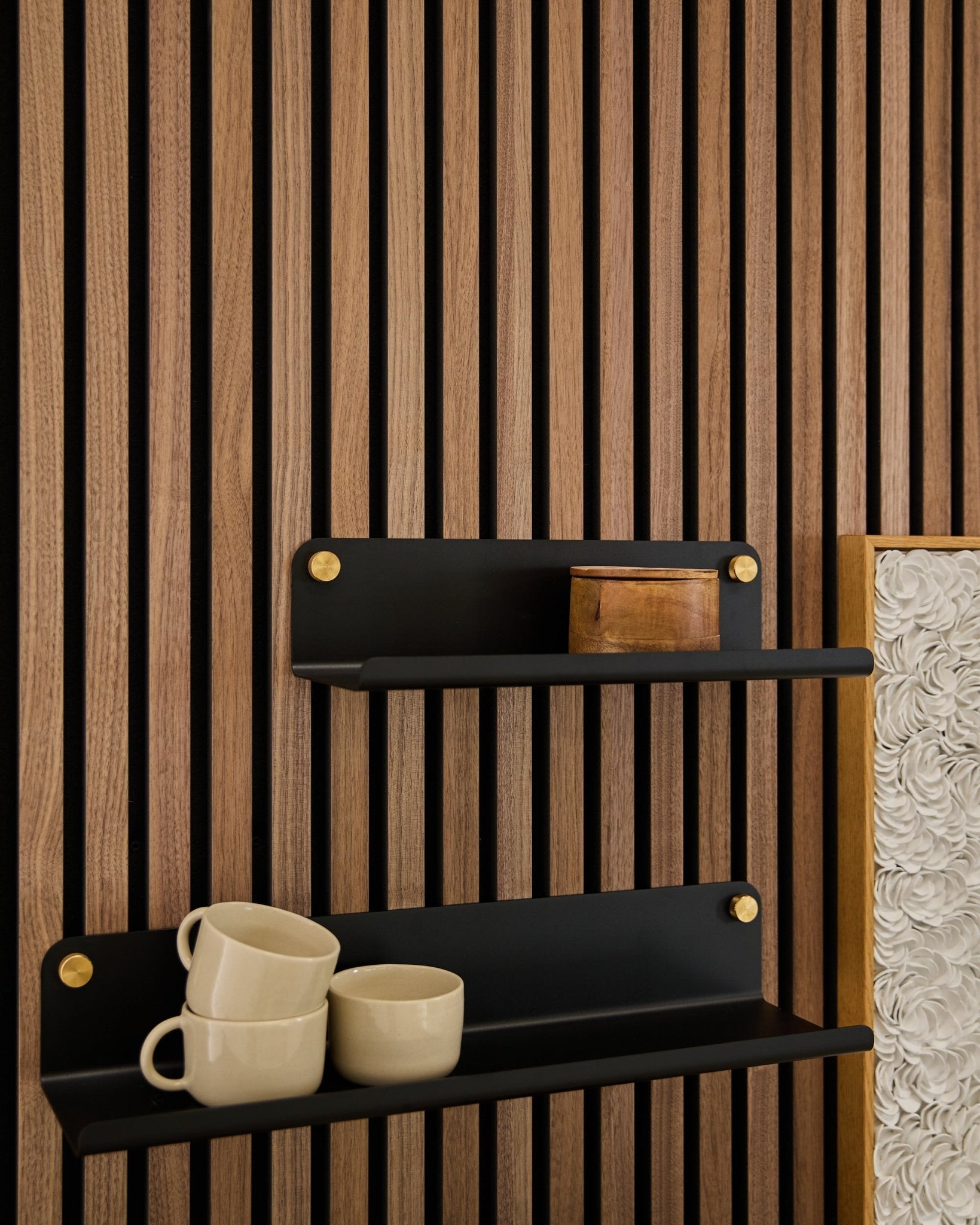 Deux étagères larges black Norr de WoodUpp (35,9 cm) avec des supports dorés, montées sur un panneau acoustique en bois Akupanel ; l’étagère du bas accueille 3 mugs beige, celle du haut une boîte en bois small.